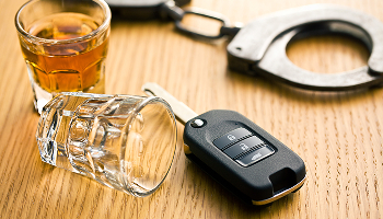 A shot glass with amber liquid, a car key, and metal handcuffs on a wooden surface representing DUI defense