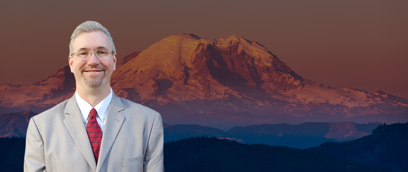 Attorney Jason Scott Newcombe in a grey suit and red tie with a scenic view of Mount Rainier at sunset in the background