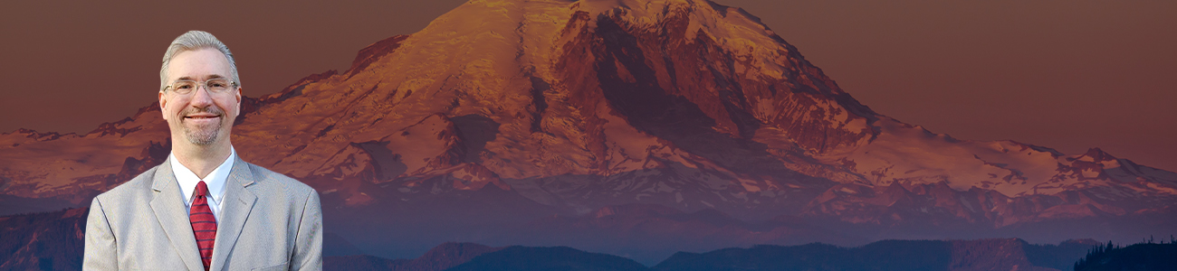 Attorney Jason Scott Newcombe in a grey suit and red tie with a scenic view of Mount Rainier at sunset in the background