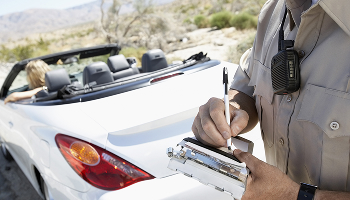 A police officer in a tan uniform writing a ticket next to a white convertible car representing t raffic ticket defense
